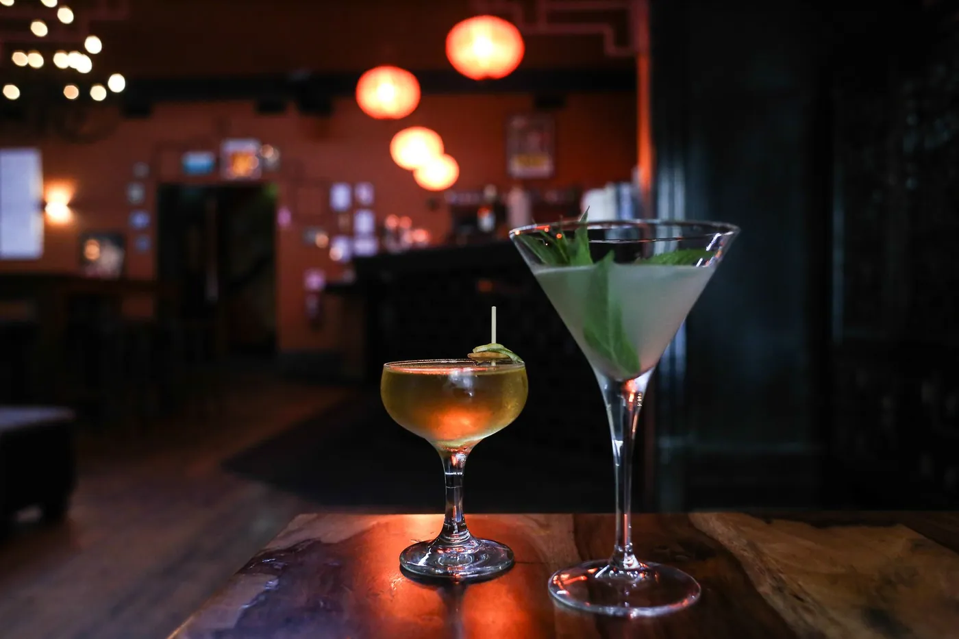 Două cocktailuri pe o masă de lemn într-un bar cu lumină scăzută și lămpi calde în fundal